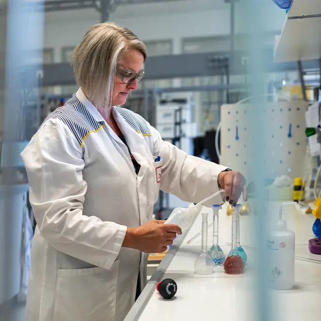 一位女性科学家穿着实验室白大褂,在实验室内进行实验。她正用吸管操作不同的试管,周围有各种实验器材和化学品。