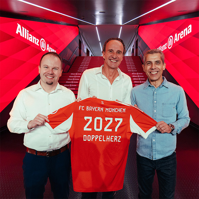 在中旗帜鲜明的安联球场，三位男士微笑着合影，手中展示一件印有“FC Bayern München 2027 Doppelherz”字样的橙色足球衫。背景为红色光辉的墙面，增添了热烈的体育氛围。