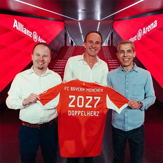 在中旗帜鲜明的安联球场,三位男士微笑着合影,手中展示一件印有“FC Bayern München 2027 Doppelherz”字样的橙色足球衫。背景为红色光辉的墙面,增添了热烈的体育氛围。
