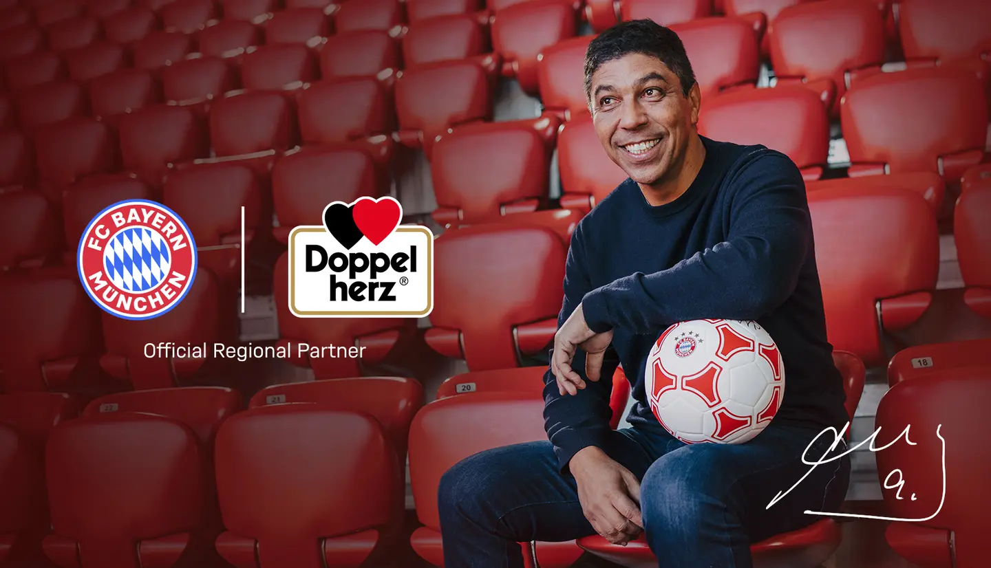 A smiling man sits in a stadium seat, holding a soccer ball. To his left, there are logos for FC Bayern München and Doppelherz, with the text "Official Regional Partner" underneath. The background features empty red seats.